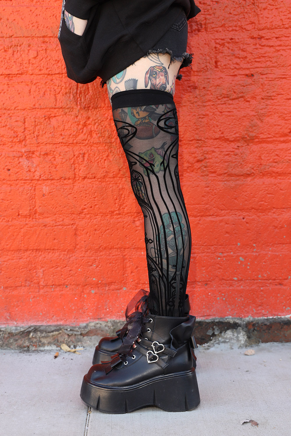 A person's lower body wearing black thigh-high sheer patterned socks or stockings with black platform lace-up boots, standing against a bright orange brick wall.
