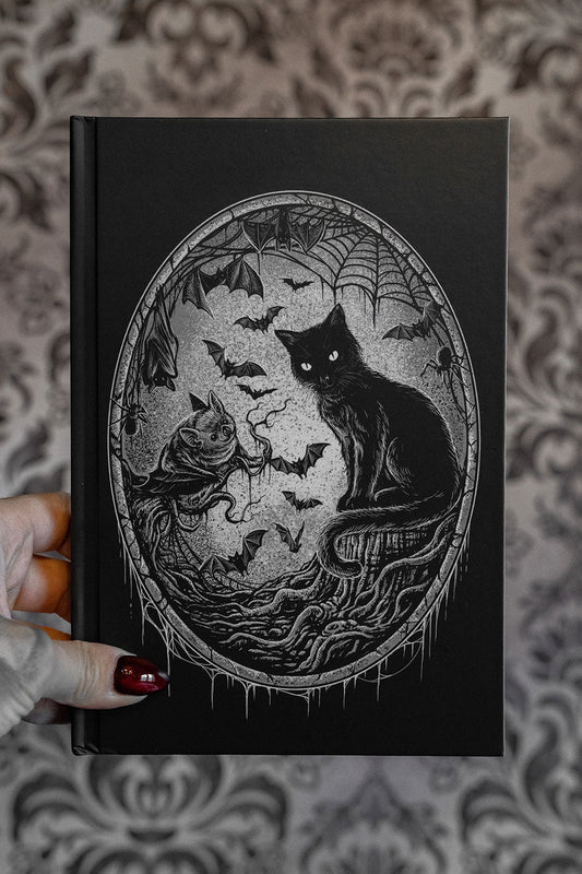 Black notebook with a cat and bats illustration held by a hand against a patterned background